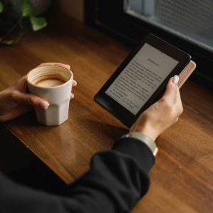 A person holding an ereader tablet drinking a coffee at a table.