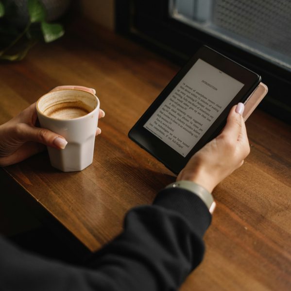 A person holding an ereader tablet drinking a coffee at a table.