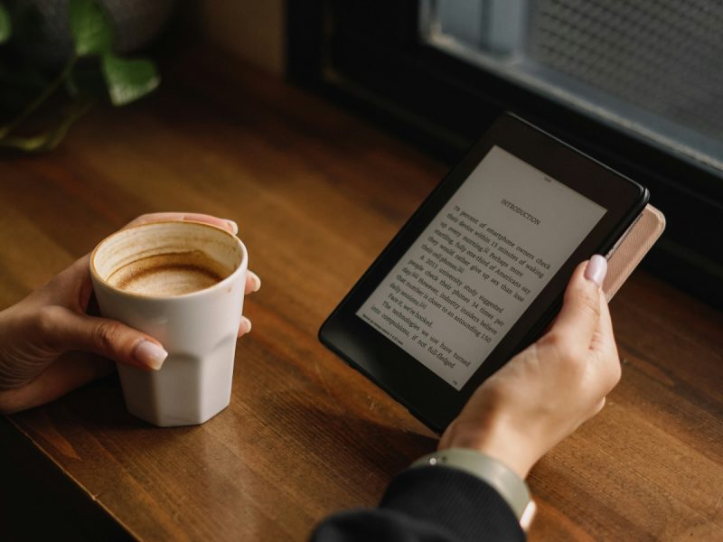 A person holding an ereader tablet drinking a coffee at a table.